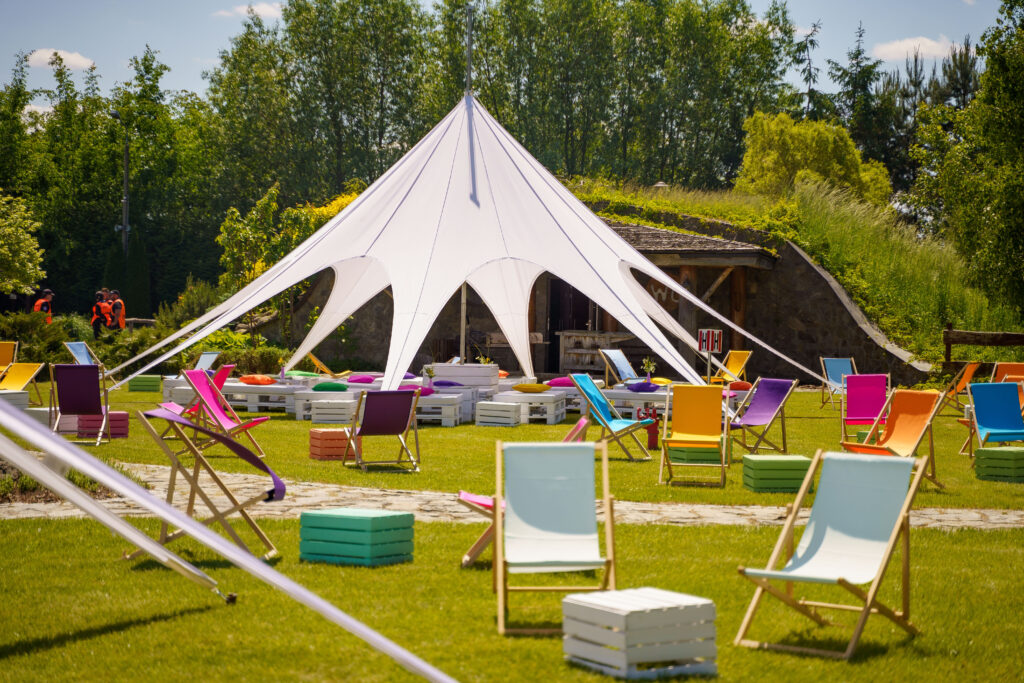 Na zdjęciu widać kolorowe leżaki oraz białą, rozstawioną płachtę namiotową w zielonym plenerze, co tworzy przyjazną atmosferę imprezy plenerowej.