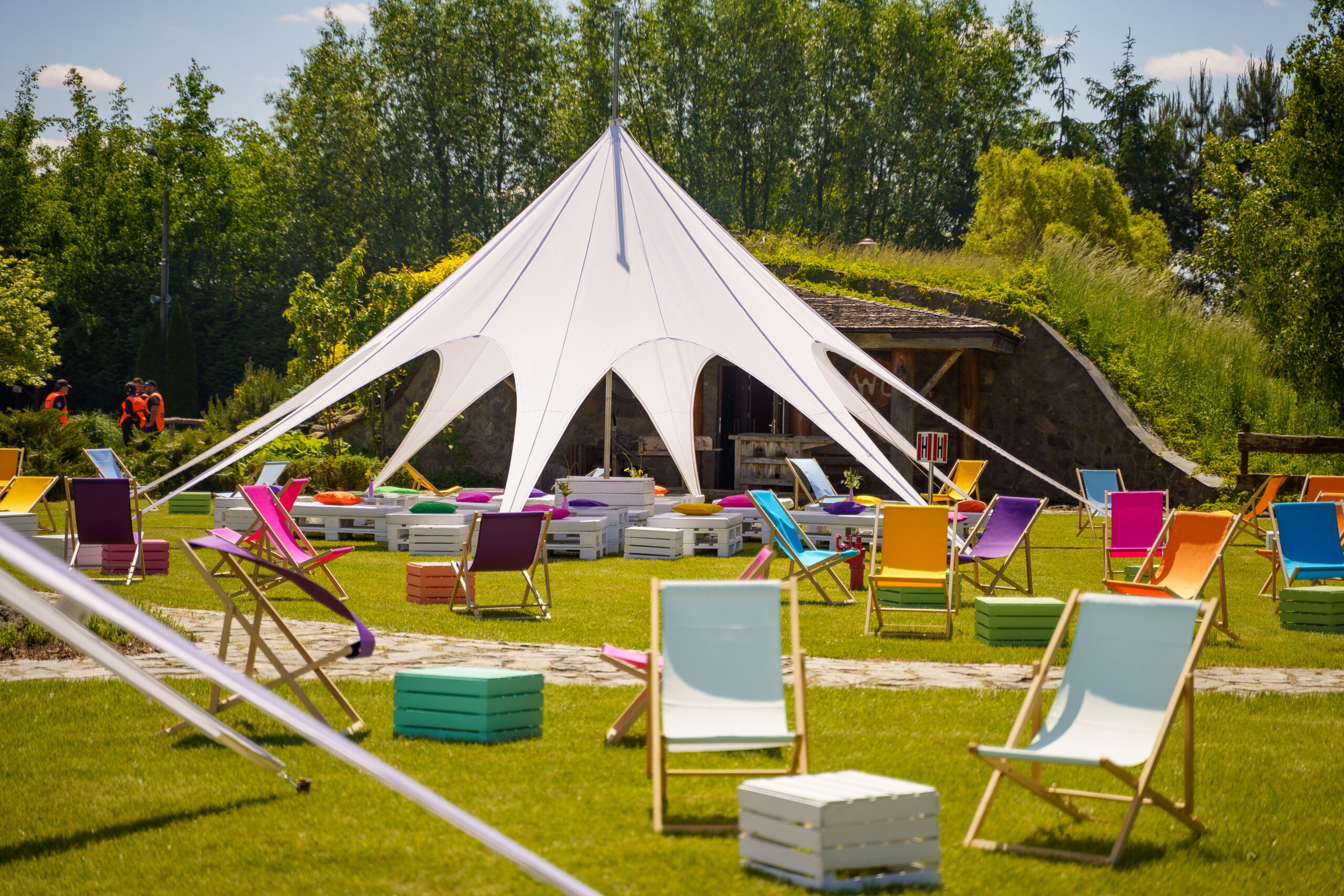 Na zdjęciu widać kolorowe leżaki oraz białą, rozstawioną płachtę namiotową w zielonym plenerze, co tworzy przyjazną atmosferę imprezy plenerowej.