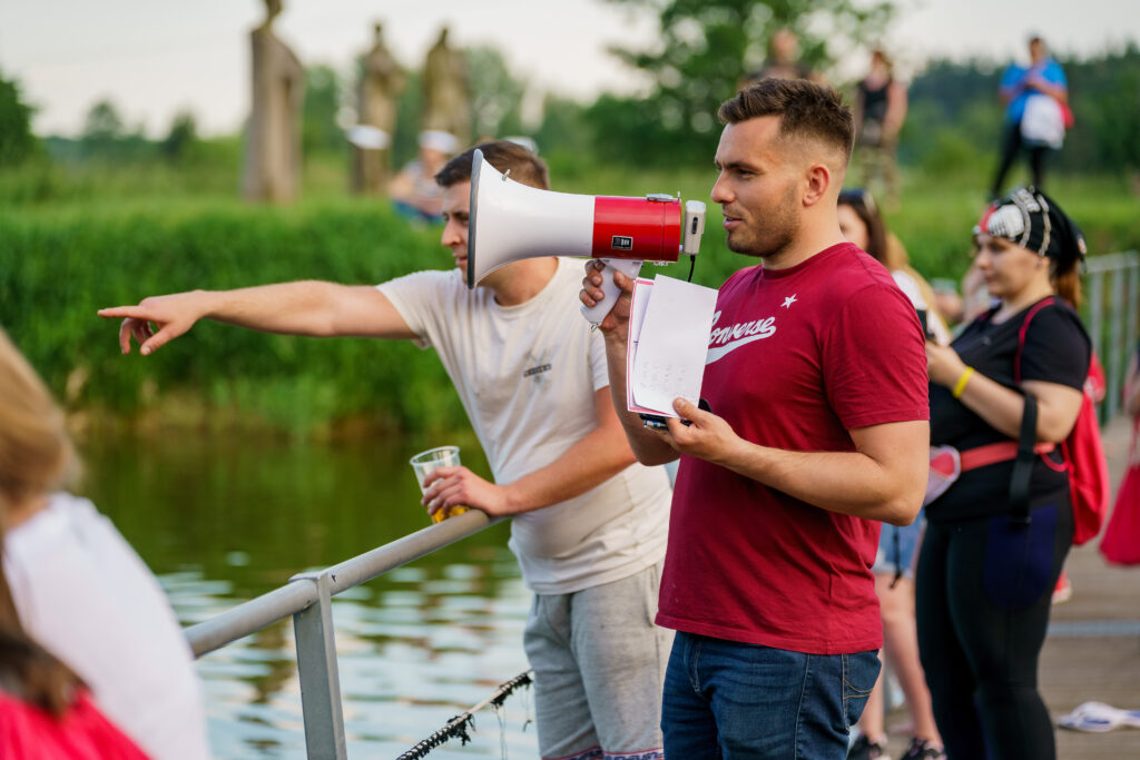 Mężczyzna trzymający megafon i kartkę z notatkami prowadzi zdarzenie na świeżym powietrzu przy wodzie, podczas gdy inna osoba pokazuje coś w oddali.