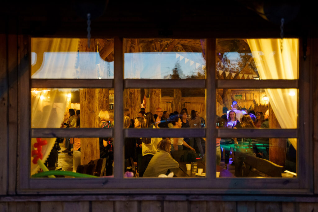 Widok przez okno na wnętrze drewnianej chaty, gdzie odbywa się impreza z udziałem licznych gości.