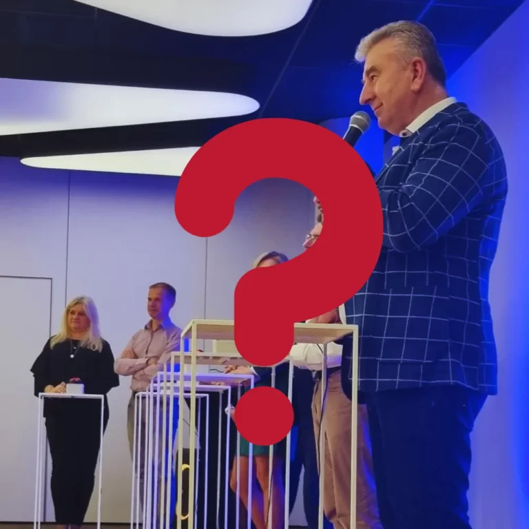 Na zdjęciu widzimy osobę przemawiającą do mikrofonu w pomieszczeniu, w którym znajdują się inni ludzie, a w centralnej części grafiki umieszczono duży czerwony znak zapytania.