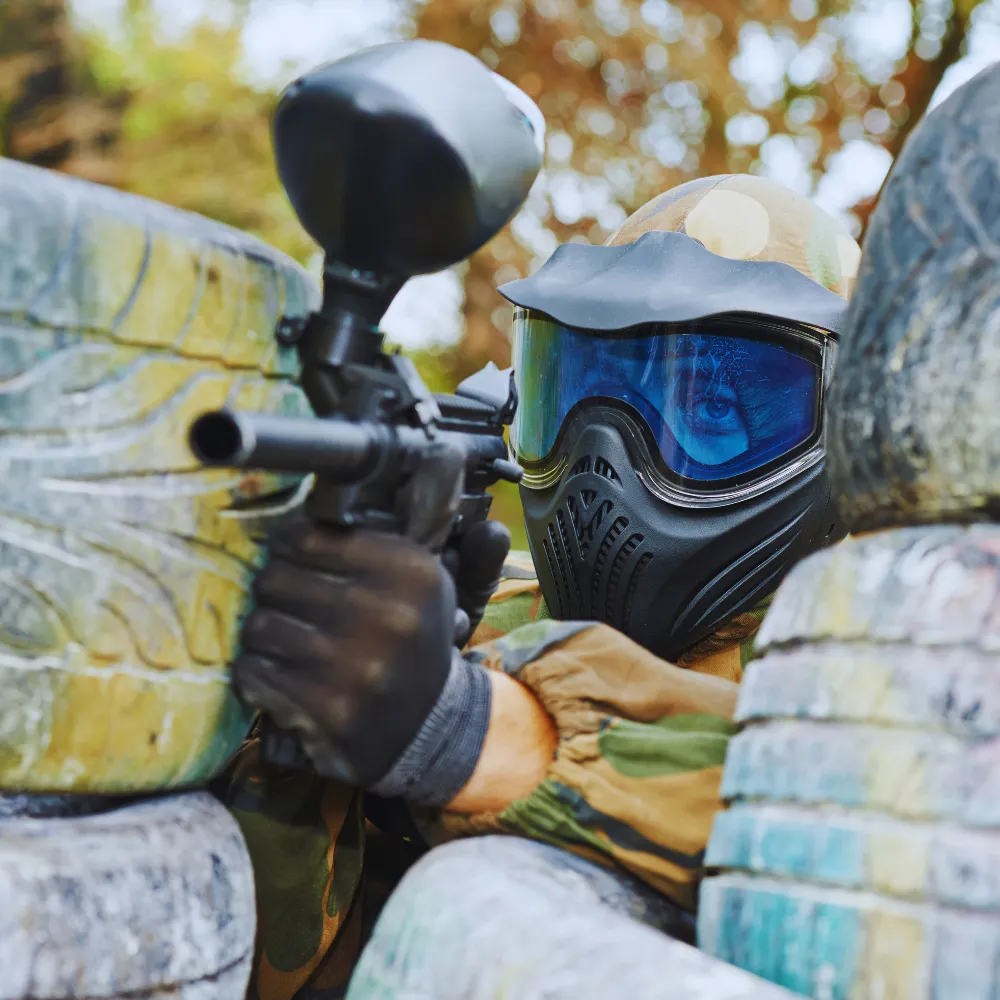 Zdjęcie przedstawia osobę w stroju do paintballa, celującą z markera zza przeszkód z opon.