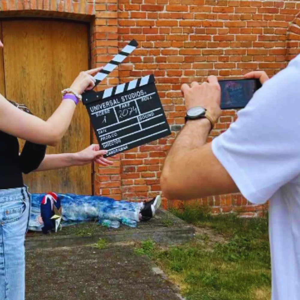 Na zdjęciu widzimy scenę filmową, gdzie jedna osoba trzyma klapsa filmowego, a druga nagrywa telefonem, z kimś leżącym na ziemi w tle.