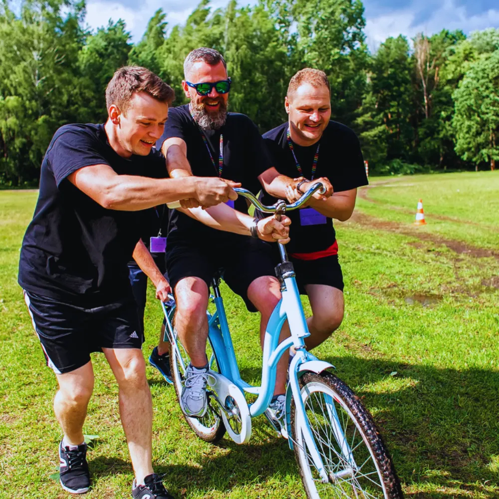 Trzech mężczyzn ubranych w czarne koszulki z uśmiechami jedzie na nietypowym rowerze typu tandem na zielonej trawie, otoczeni drzewami.