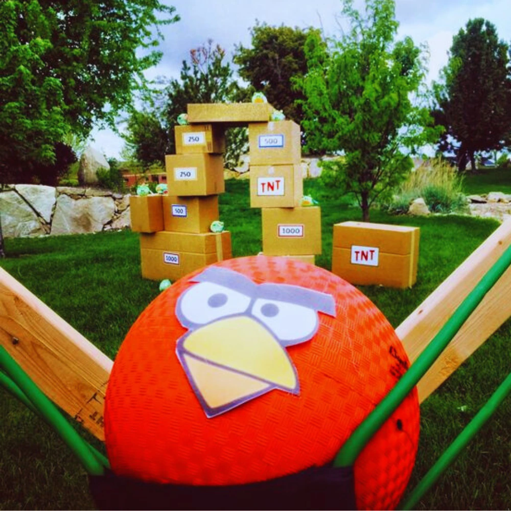 Gra plenerowa inspirowana Angry Birds z dużą katapultą i ustawionymi kartonowymi pudłami z punktami i oznaczeniem TNT na trawniku.