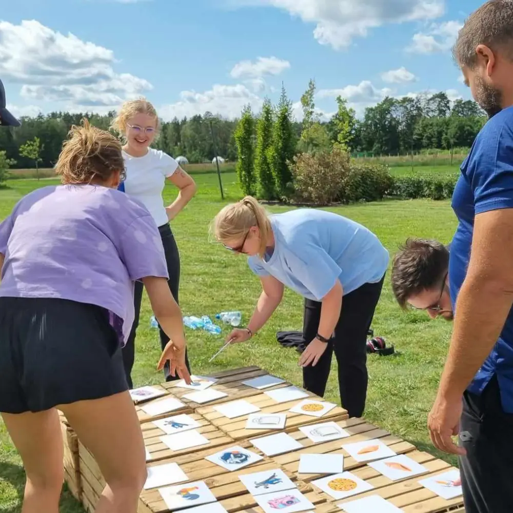 Grupa ludzi bierze udział w aktywności na świeżym powietrzu, analizując karty ułożone na drewnianej platformie na tle zielonego terenu.
