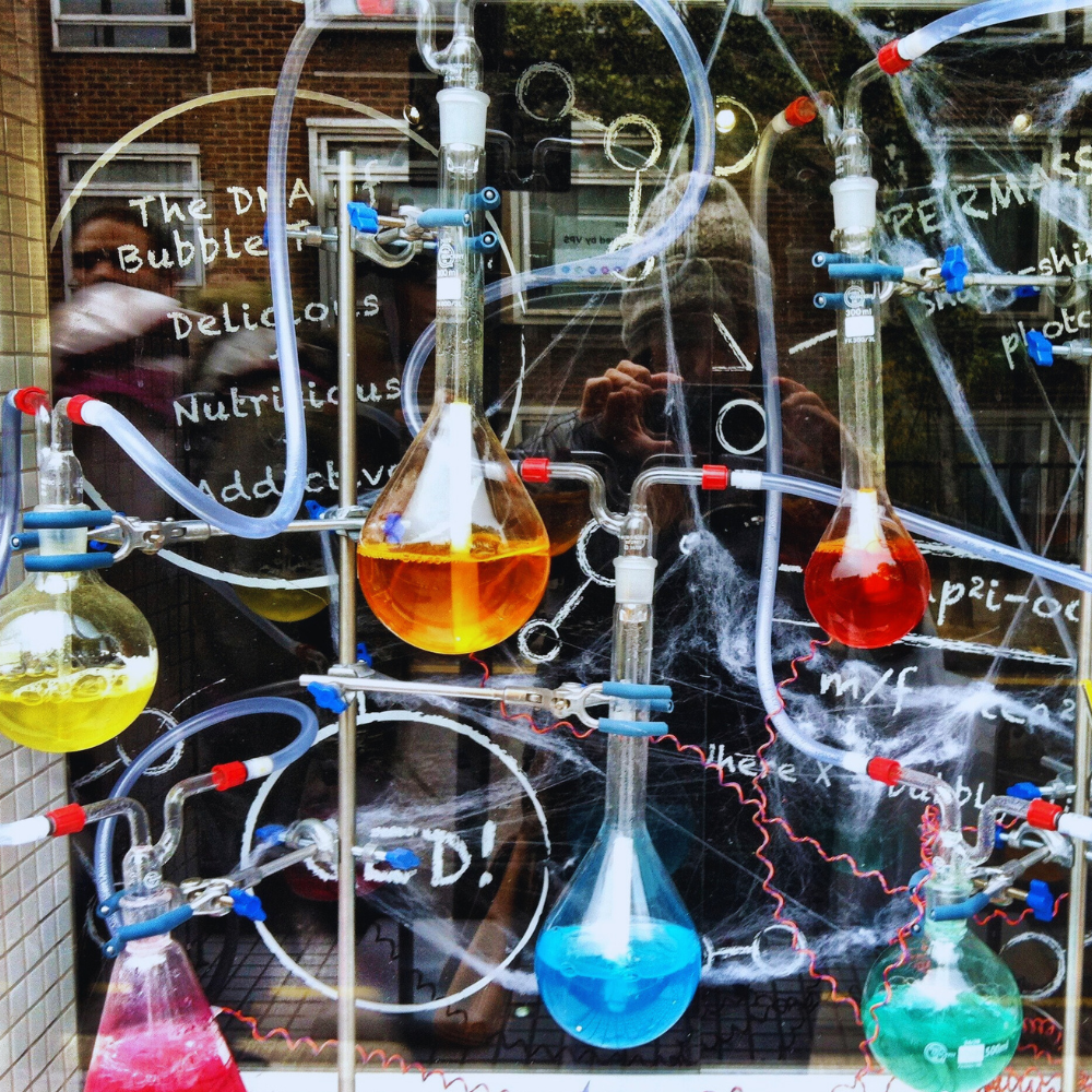 Kolorowy układ chemiczny z fiolkami i rurkami pełnymi cieczy na tle szkoły, przypominający dekorację eventową.